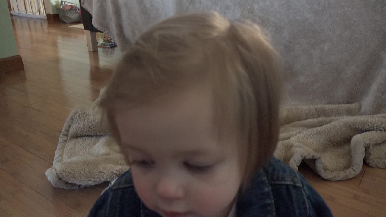 The toddler remains close to the lens in the Westminster home, looking downward with the gray couch and folded blanket behind her.