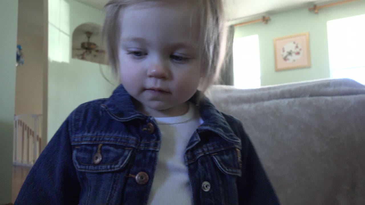 Blonde toddler in a dark denim jacket standing close to a low indoor camera.