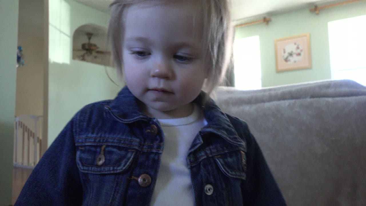 At 0:01 in Westminster, Colorado, the toddler remains close to the camera, looking down with pale green walls, a sofa back, windows, and framed art in the background.
