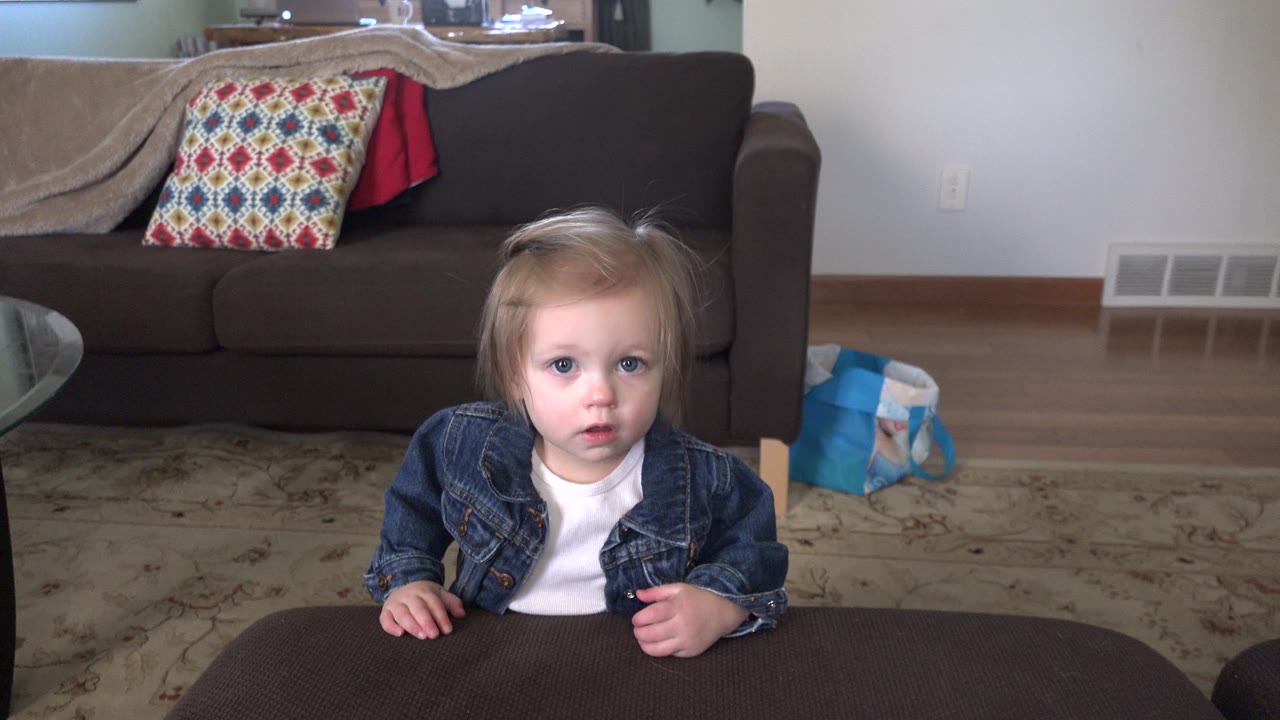 Toddler in denim jacket at an ottoman in a living room.