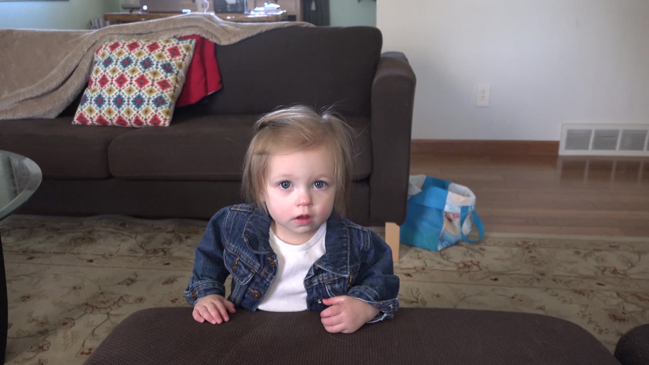The toddler remains centered at the ottoman in the Westminster, Colorado living room, facing the camera with the couch, pillow, rug, and blue bag visible around them.