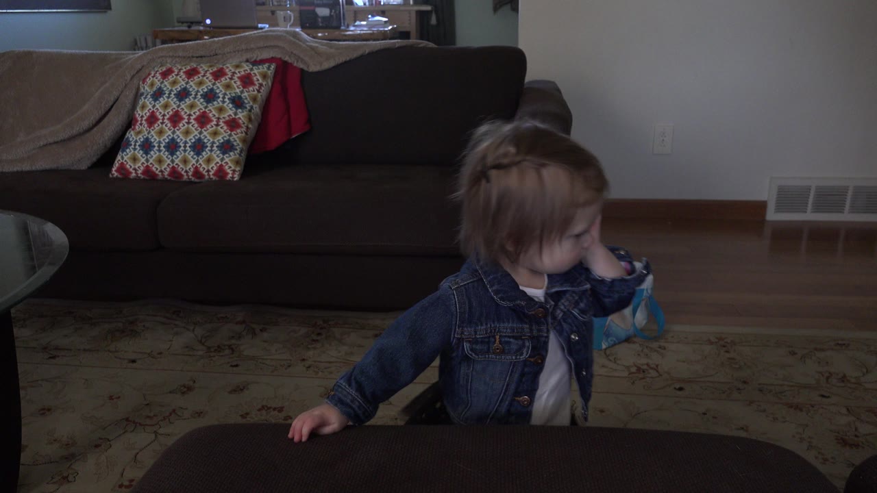 A toddler in a denim jacket stands beside a dark foreground table in a Westminster, Colorado living room with a couch and patterned pillow behind them.