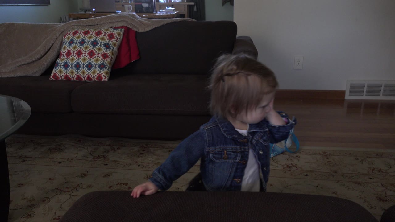 The child remains near the table edge in the Westminster home interior, turned sideways with one hand raised near their face and the couch behind them.