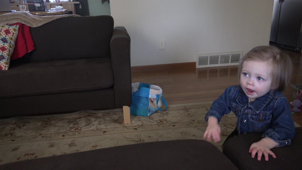 A young girl in a denim top stands at the right side of the frame by a dark ottoman in a Westminster, Colorado living room with sofa and floor vent behind her.