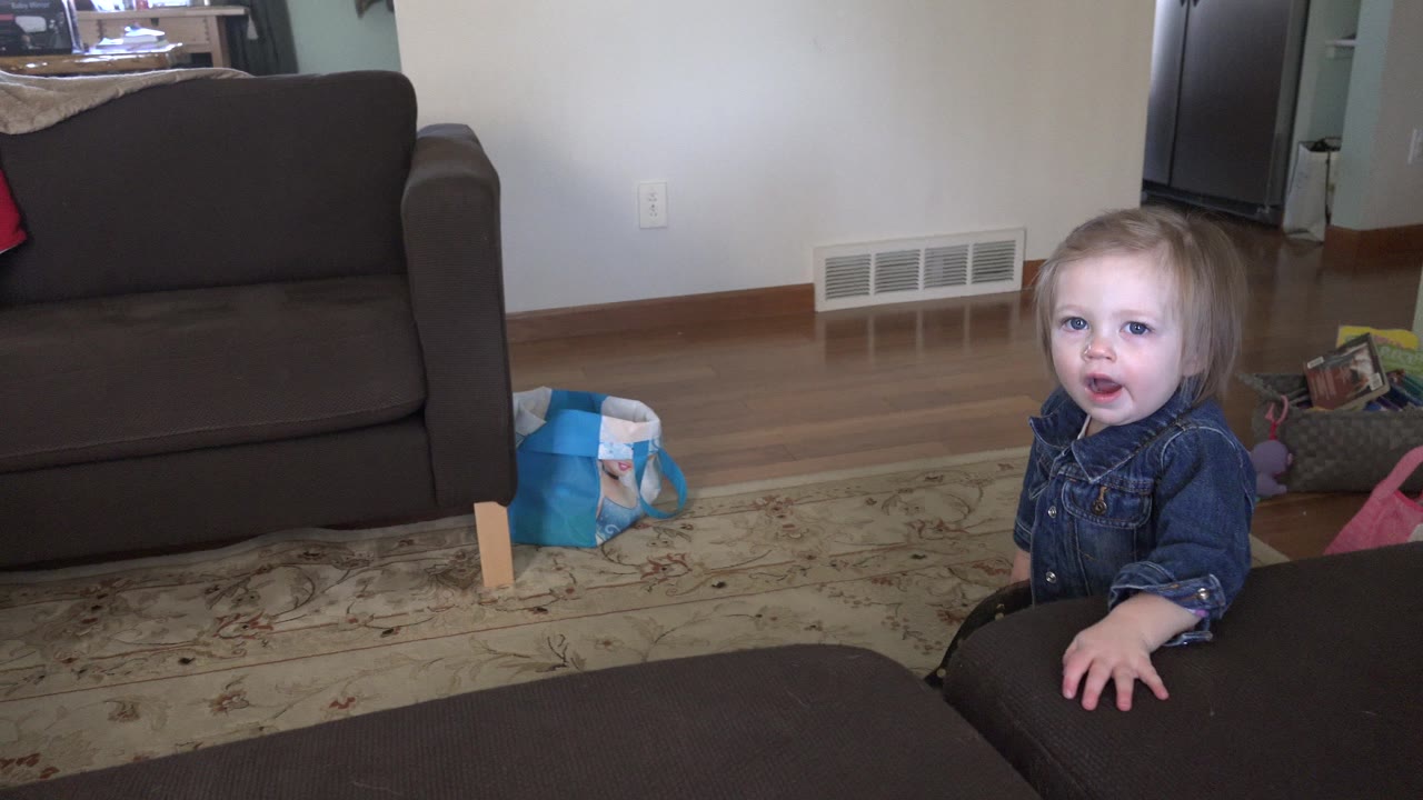 Young girl faces the camera beside a dark ottoman in a living room.