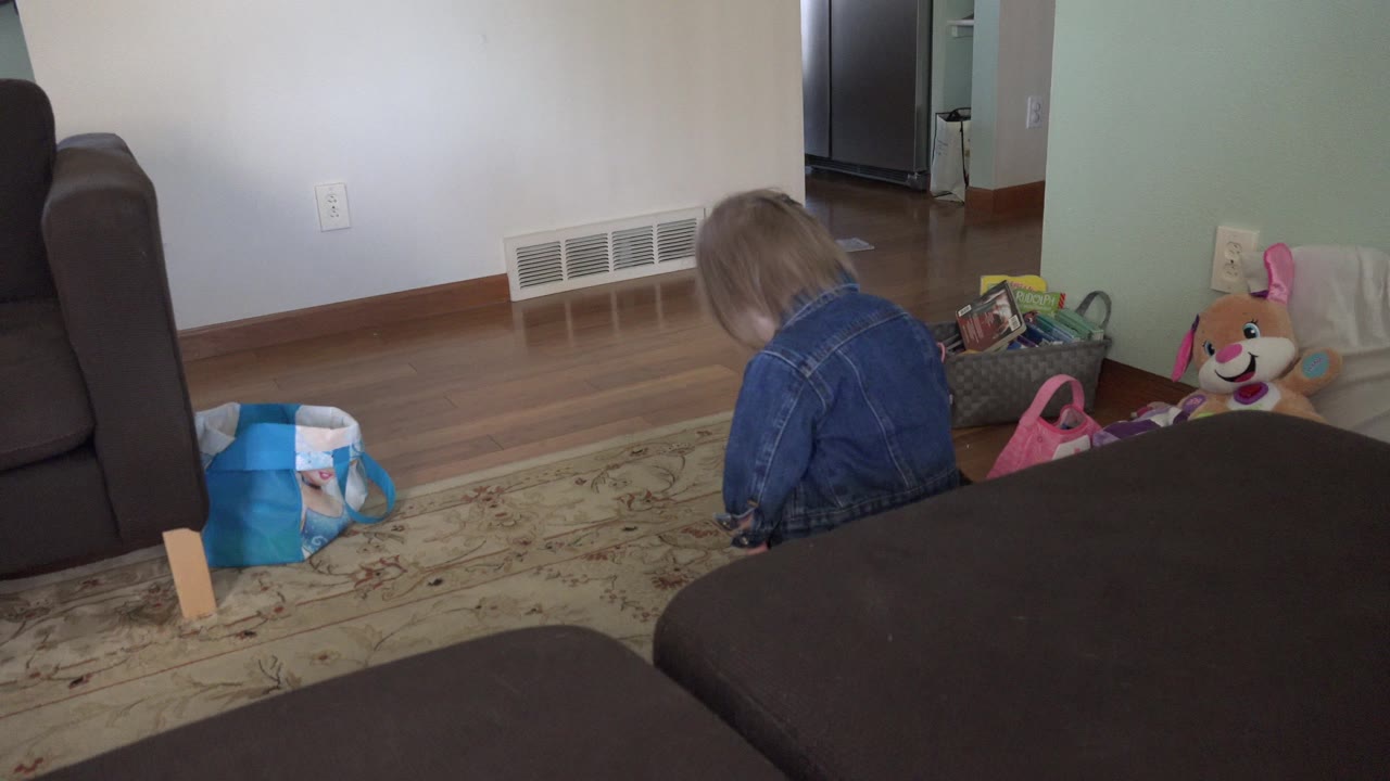 The toddler remains near the toy area on the living room floor, partly turned away from the camera, with a stuffed toy and basket visible to the right.