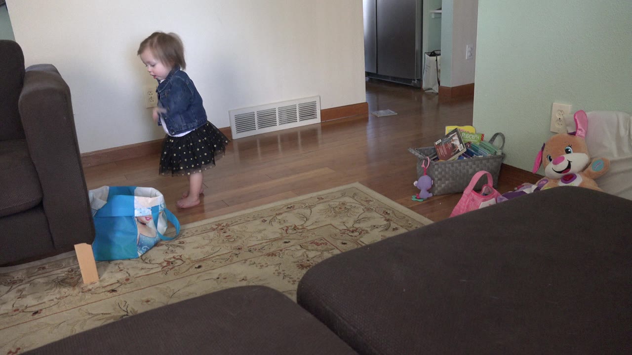 The child remains beside the wall in the Westminster, Colorado home interior, with a toy basket, stuffed toy, rug, sofa, and hallway area visible around her.