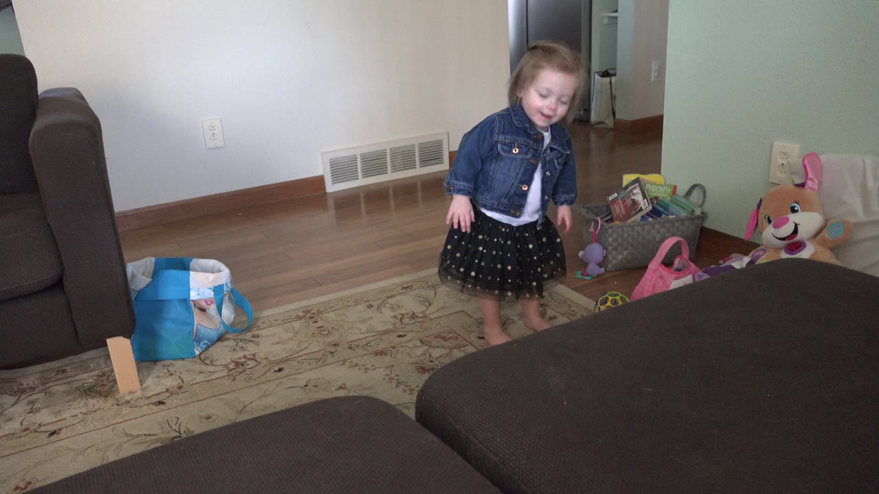 A toddler girl stands barefoot in a Westminster, Colorado living room beside toy bags, a plush dog, a sofa, a rug, and wood flooring.