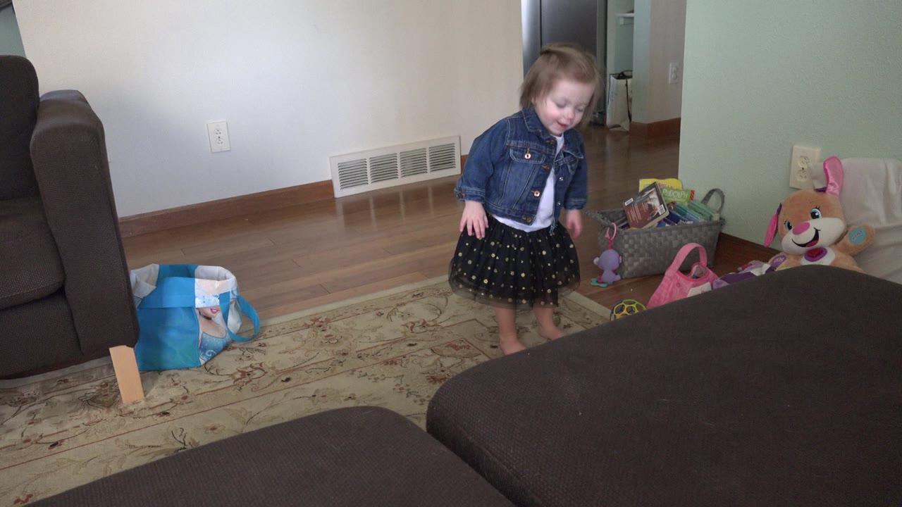 The child remains near the toy area in the Westminster living room, looking down with the sofa arm and dark ottoman framing the foreground.