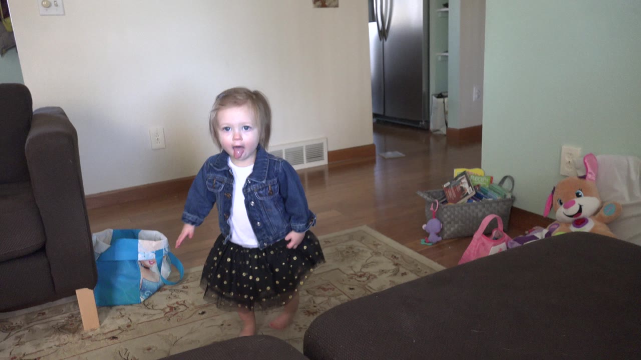The toddler girl walks toward the camera in the Westminster home interior, with the grey couch framing the left side and toys and bags visible near the wall.