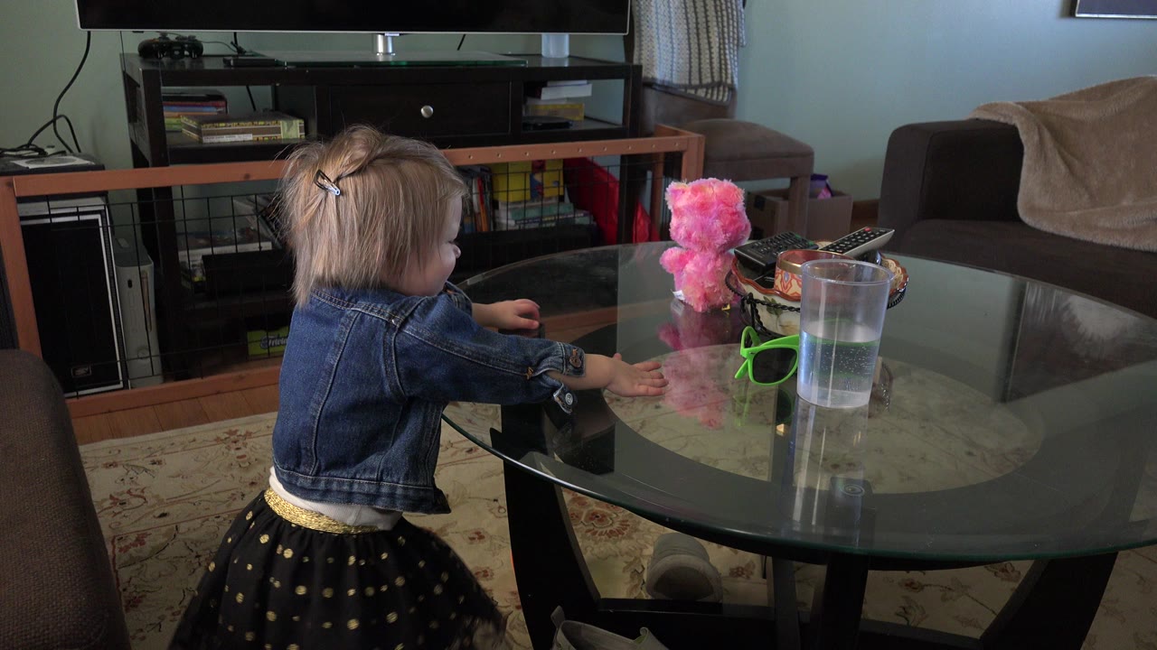 A young child in a denim jacket and dark skirt stands beside a glass coffee table in a Westminster, Colorado living room, with a pink plush toy and cup on the table.