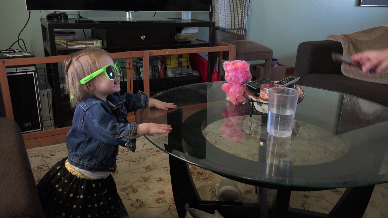 Toddler in green sunglasses reaches toward a glass coffee table.