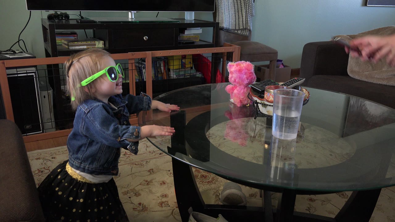 The toddler in green sunglasses looks toward an adult hand entering from the right side of the Westminster, Colorado living room frame.