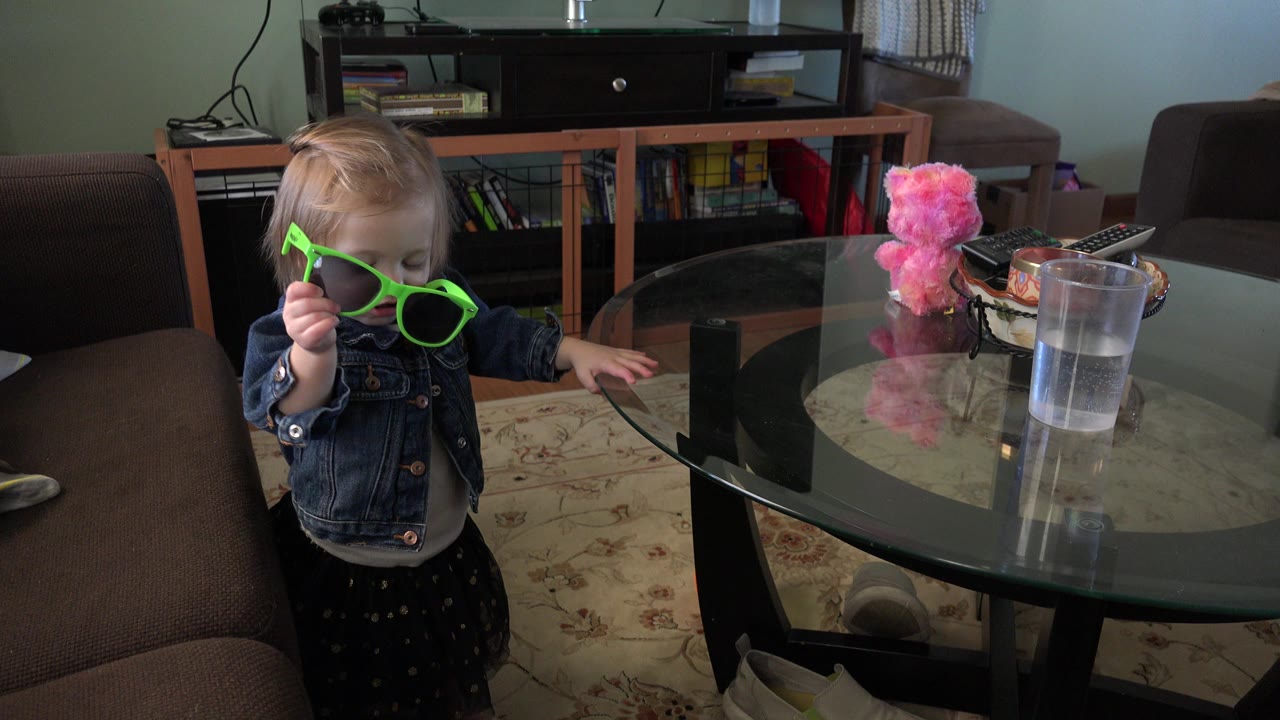 Young girl holding green sunglasses beside a glass table.