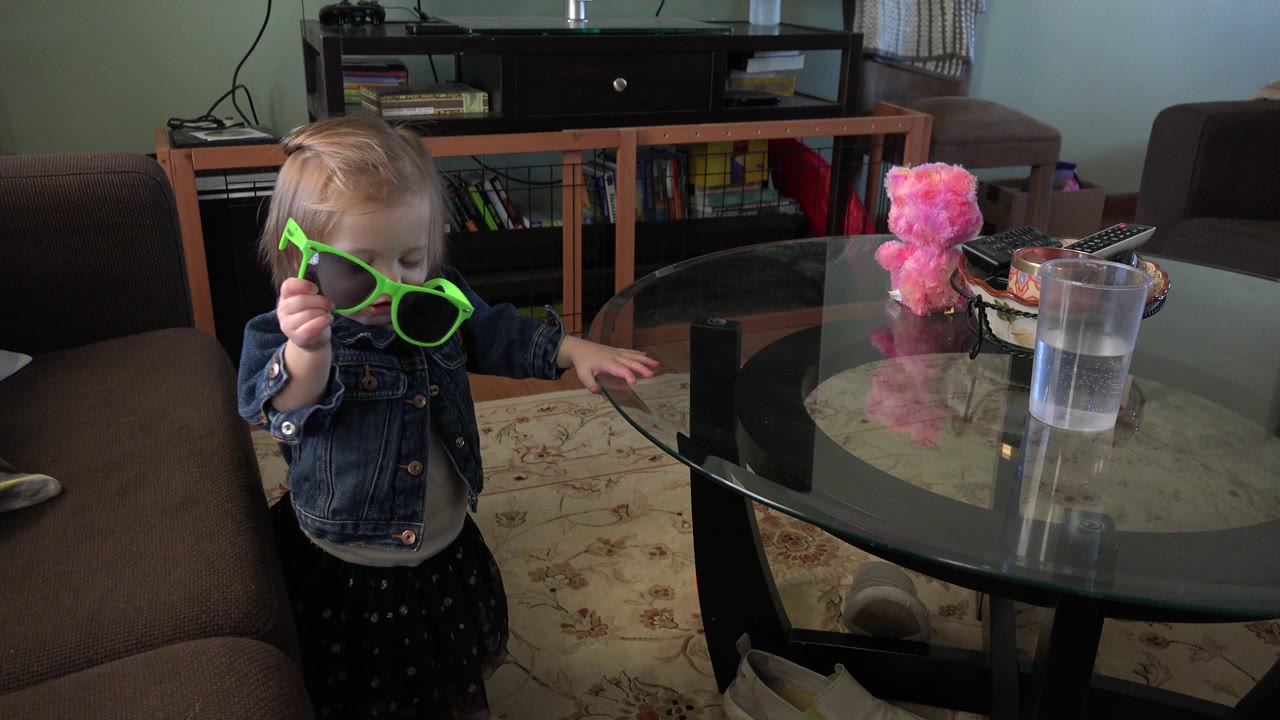 The Westminster, Colorado home frame shows the girl still holding the neon green sunglasses up, with the glass table and plastic cup prominent beside her.