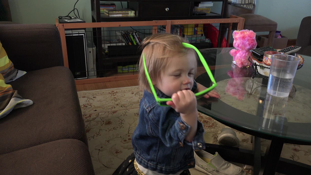 The young girl keeps the bright green sunglasses close to her mouth while standing next to the glass table in the Westminster, Colorado home interior.