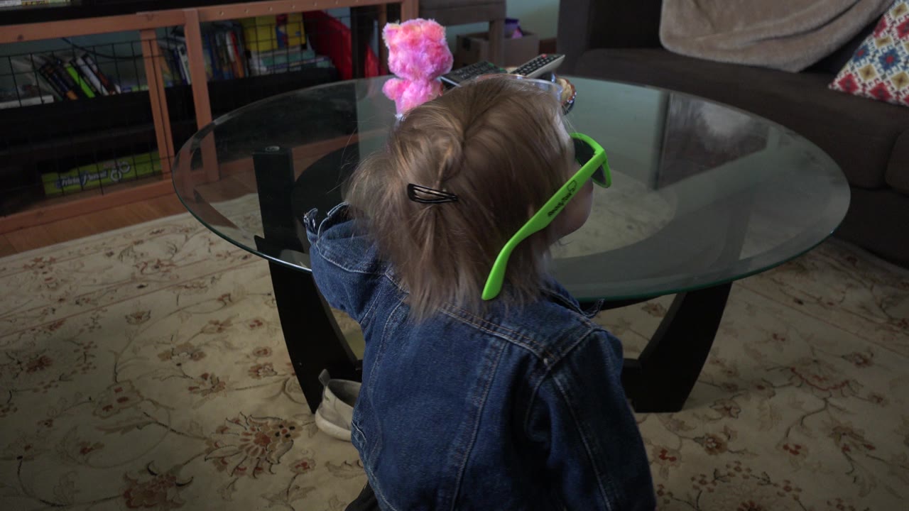 The child remains near the glass coffee table, with green sunglasses wrapping around the head and a couch, pillows, bookshelf, and pink object behind the table in Westminster, Colorado.