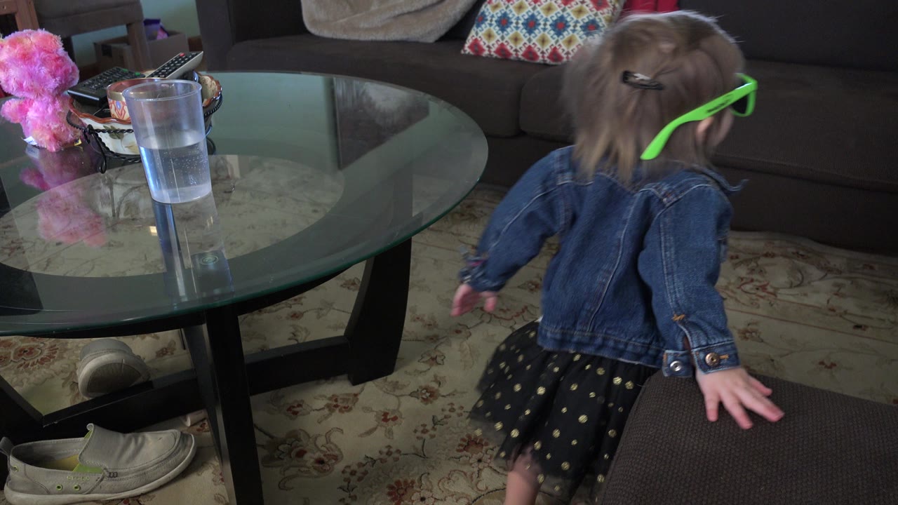 The toddler in green glasses faces toward the couch area, with one hand near a brown ottoman and the glass coffee table, water cup, and patterned pillow still visible.