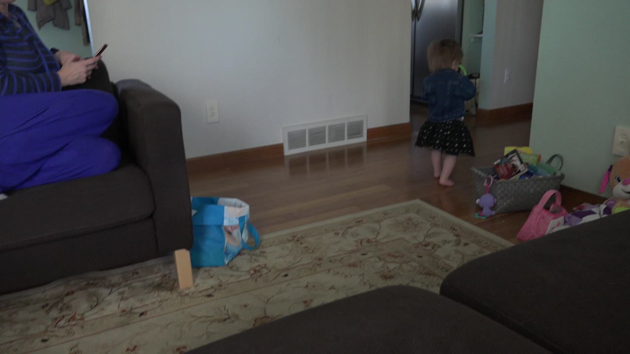 In a Westminster, Colorado living room, a toddler stands near a doorway while a partially visible adult sits on a couch holding a phone, with toys and a rug in the foreground.
