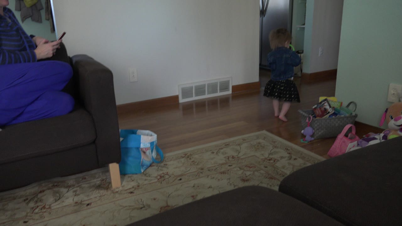 In the Westminster, Colorado home interior, the toddler faces into the doorway with toys on the floor, a couch edge at left, wall vents, and hardwood flooring visible.