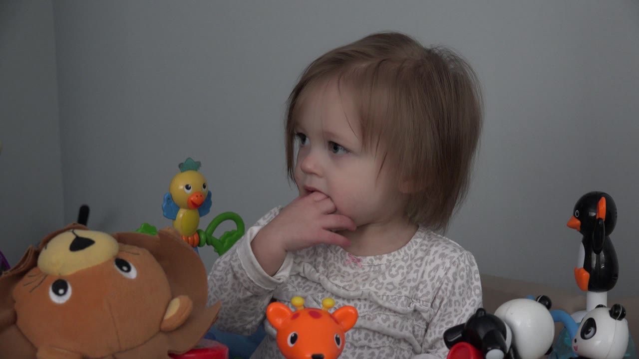The child remains seated among a plush monkey, orange toy, panda figures, and penguin toy, with her fingers in her mouth and her gaze turned left.