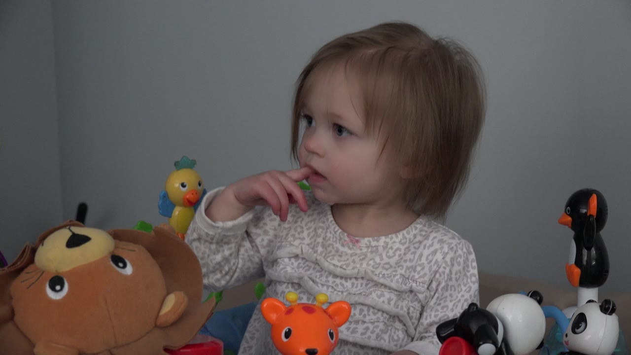 Baby in a walker with fingers near mouth among animal toys.