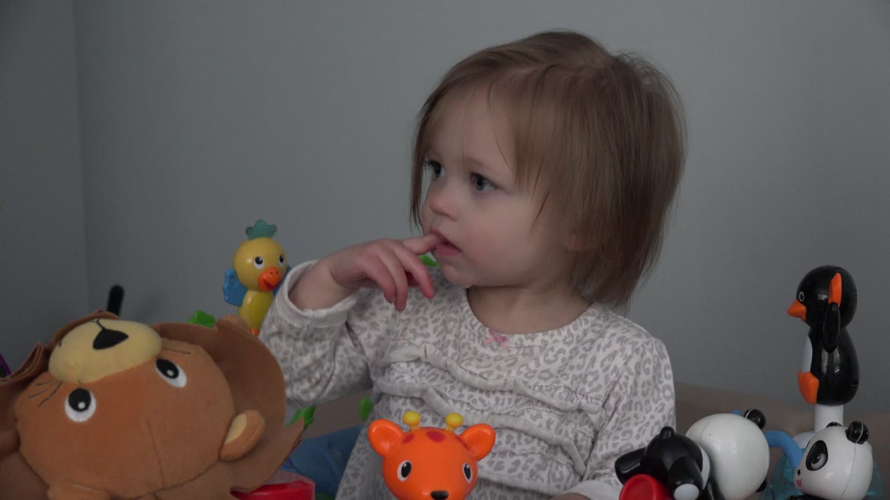 At 0:01, the baby keeps her fingers near her mouth while looking left, with the plush monkey low in front and small panda and penguin toys on the right.