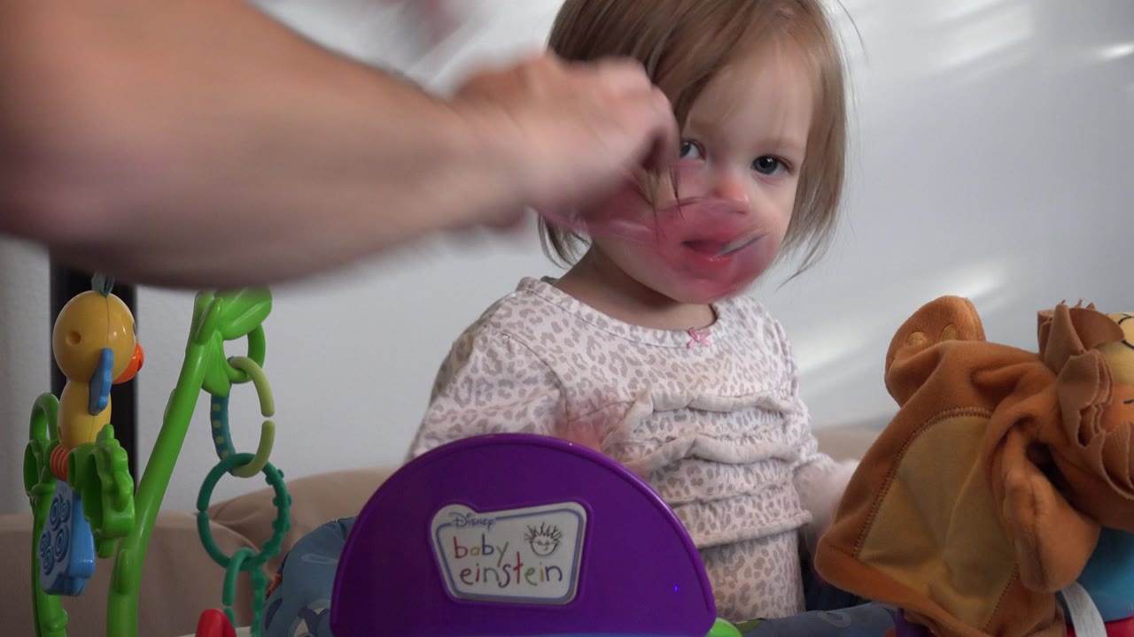 Adult hands hold pink glasses in front of a young girl seated in a toy activity seat.