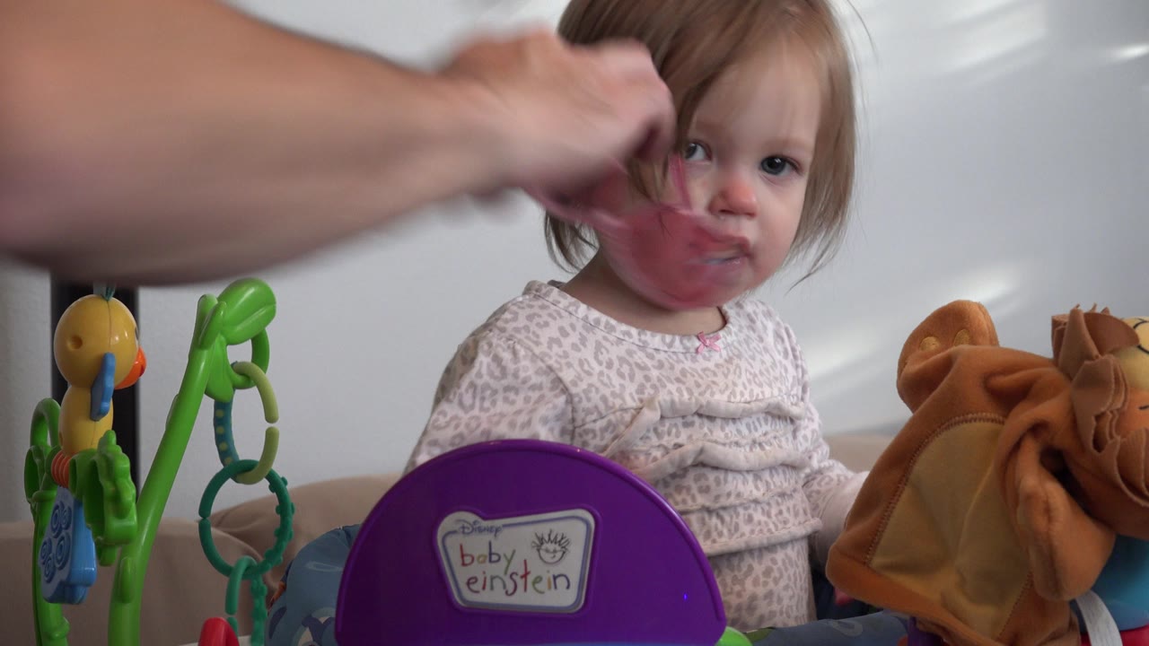 In Westminster, Colorado, the toddler looks up as adult hands hold pink glasses against the lower part of her face, with hanging toys and the purple Baby Einstein label in front.