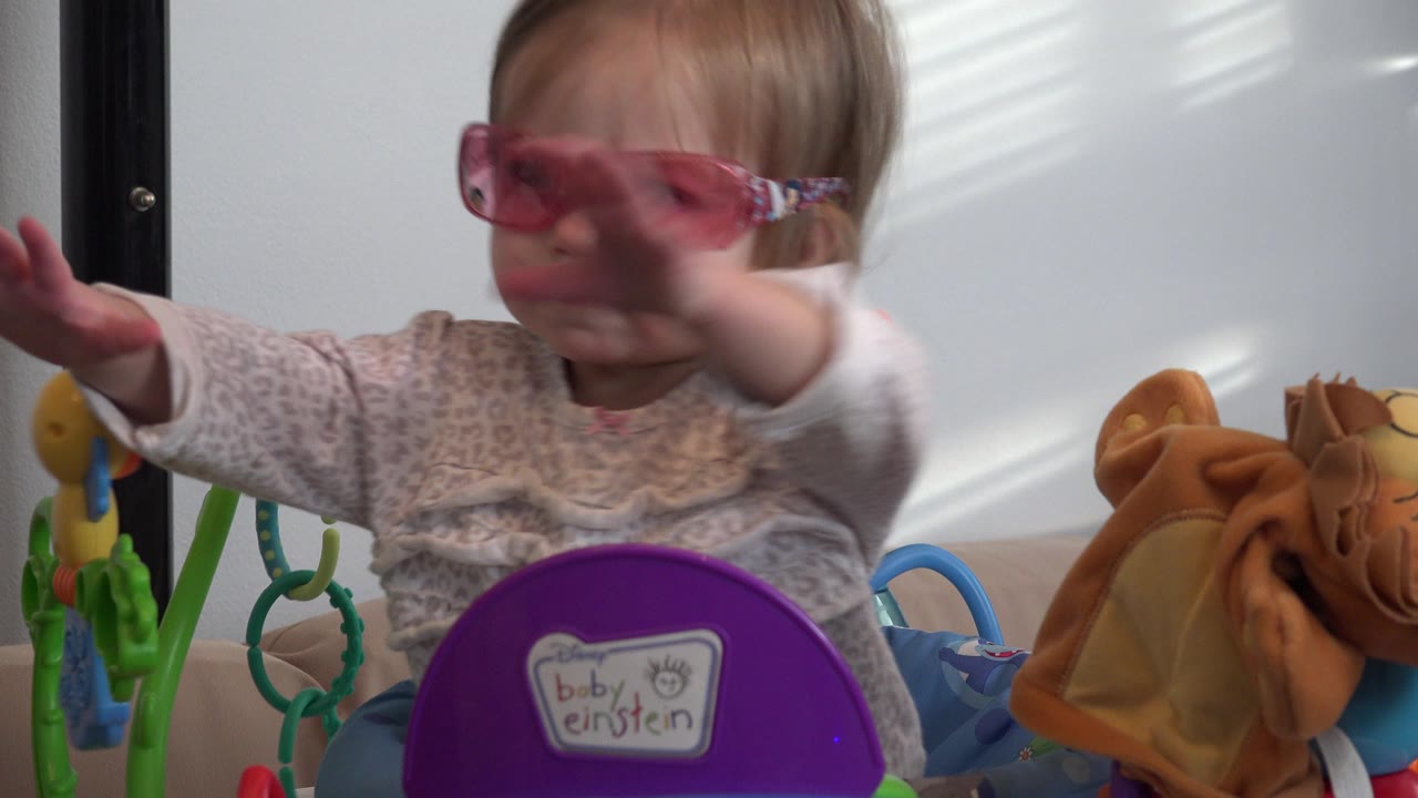 Baby girl in pink sunglasses reaches forward while seated among colorful toys.