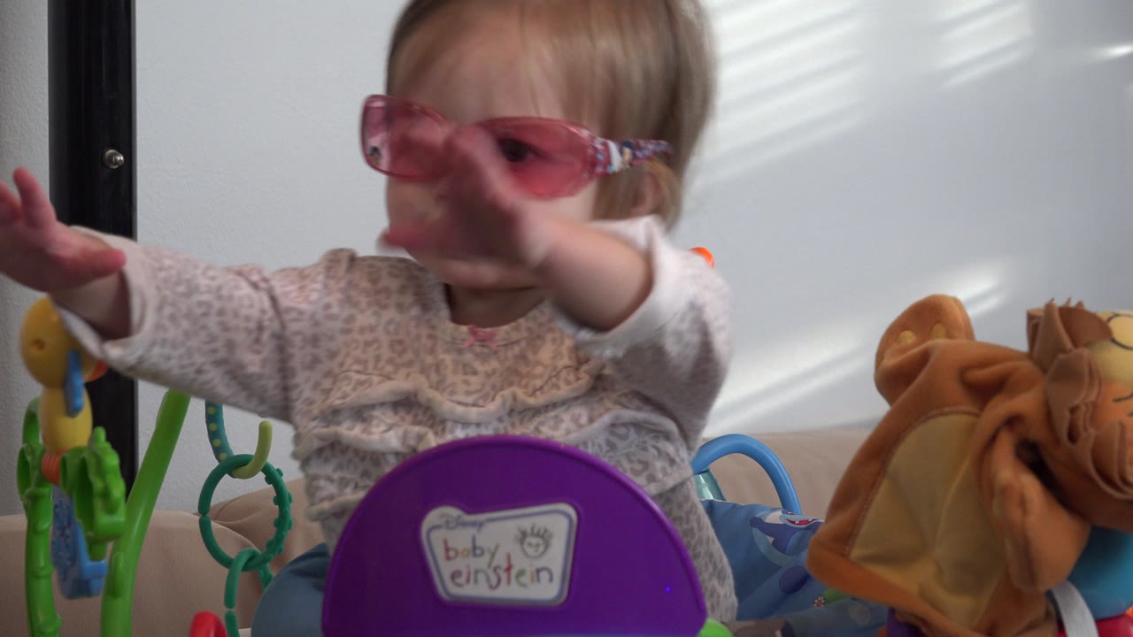 The baby girl keeps her arms extended in front of a purple Baby Einstein toy, with green play pieces and a tan plush toy around her in Westminster, Colorado.