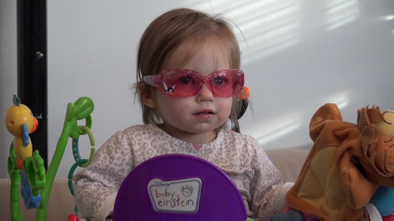 The child remains centered in Westminster, Colorado, looking forward through pink sunglasses with green and yellow toys visible on the left and a tan plush item on the right.