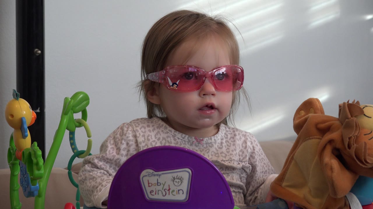 At 0:01, the toddler remains framed close in the Westminster home interior, looking forward through pink lenses with the purple toy panel low in front of her.