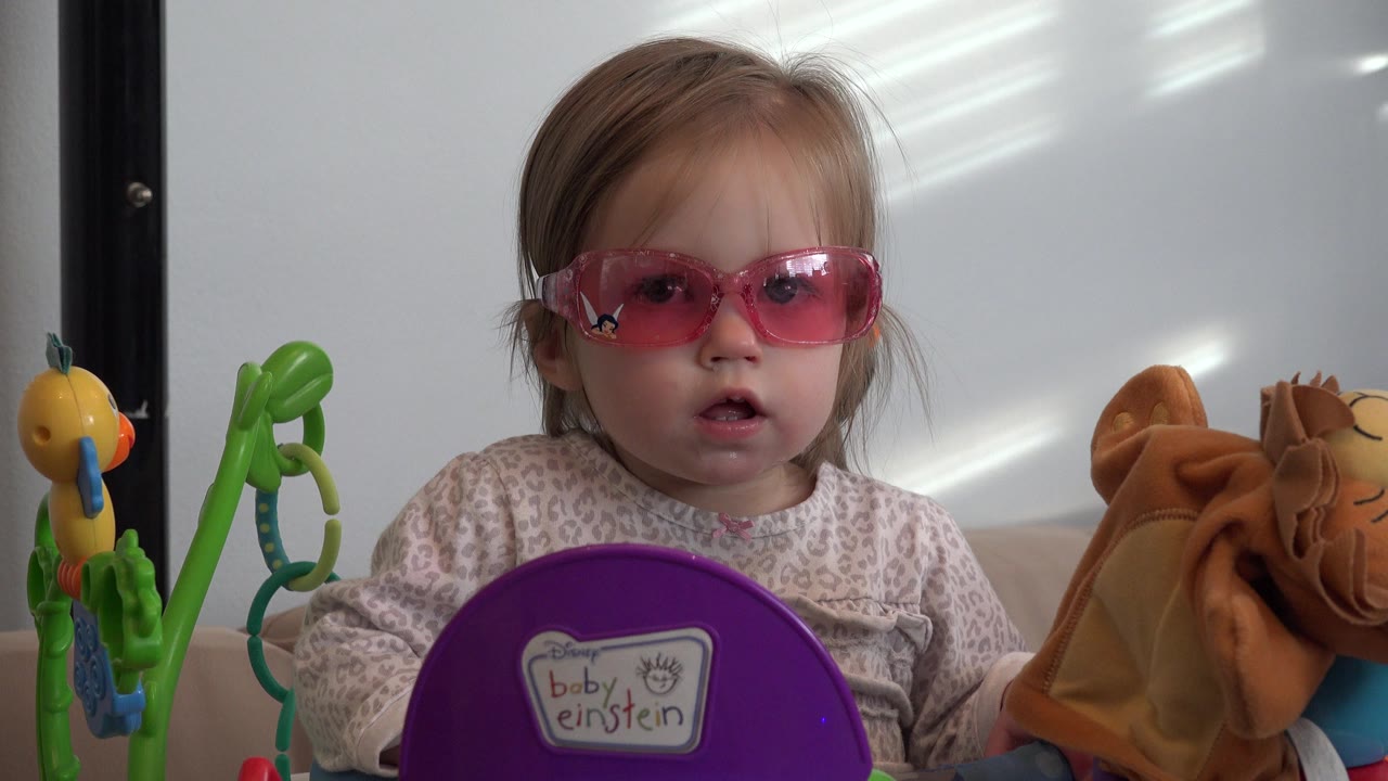 Blond baby girl wearing pink sunglasses sits among toys indoors.