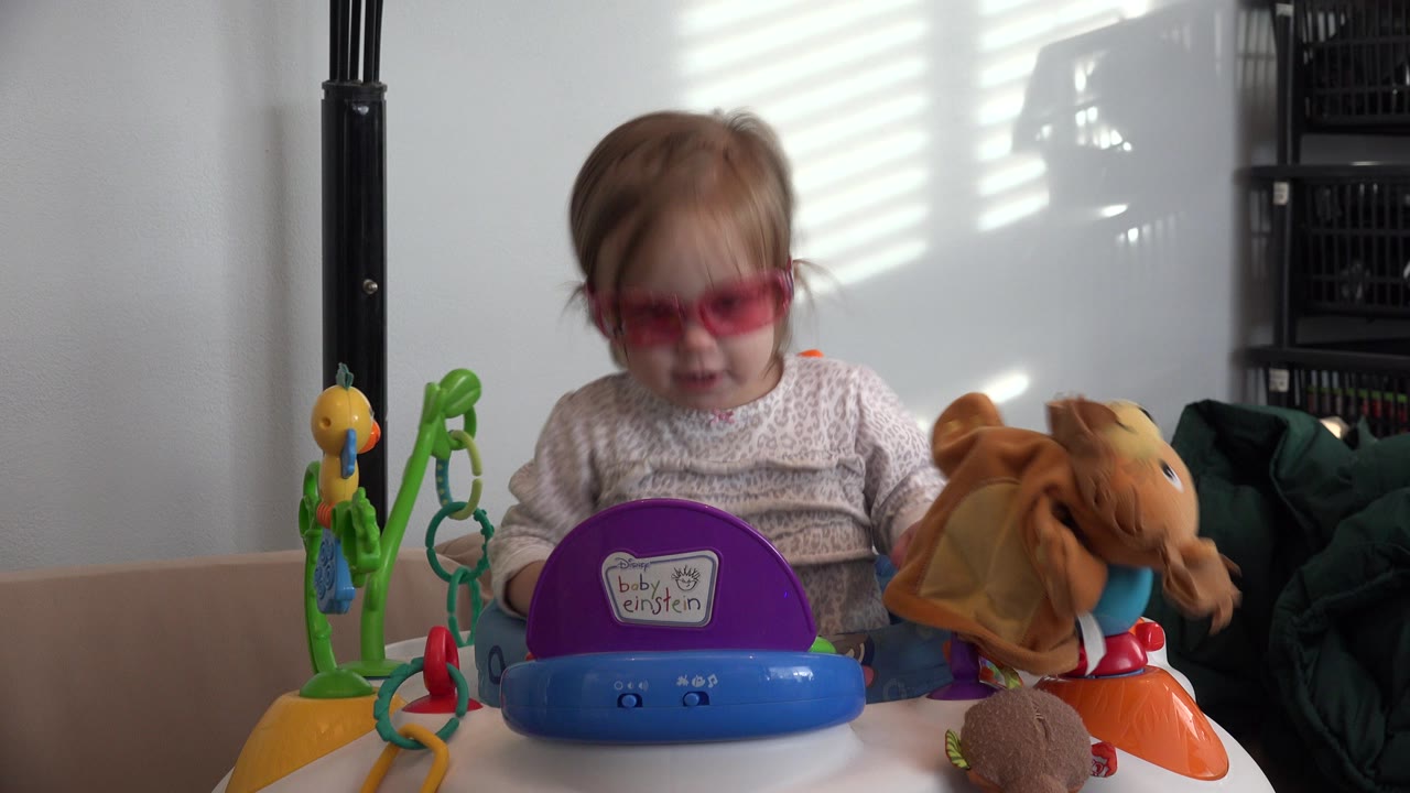 A young child in pink tinted glasses appears behind colorful stroller toys in Westminster, Colorado, with striped window light on the wall.
