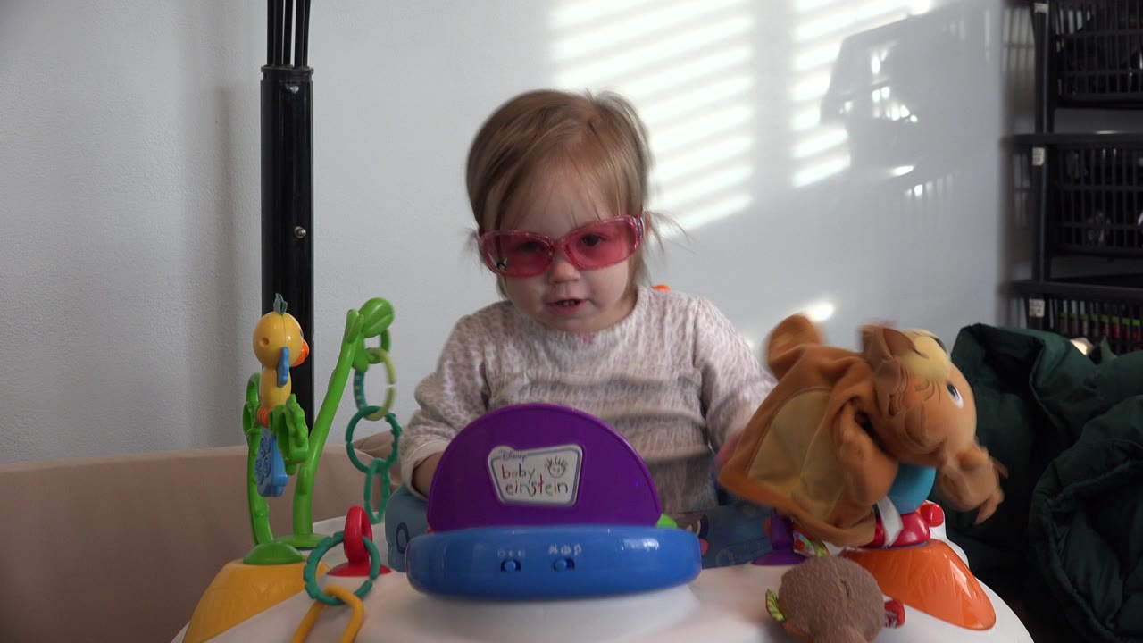 The child in pink glasses looks downward behind the purple and blue stroller toy panel, with a plush animal visible at the right edge in Westminster, Colorado.