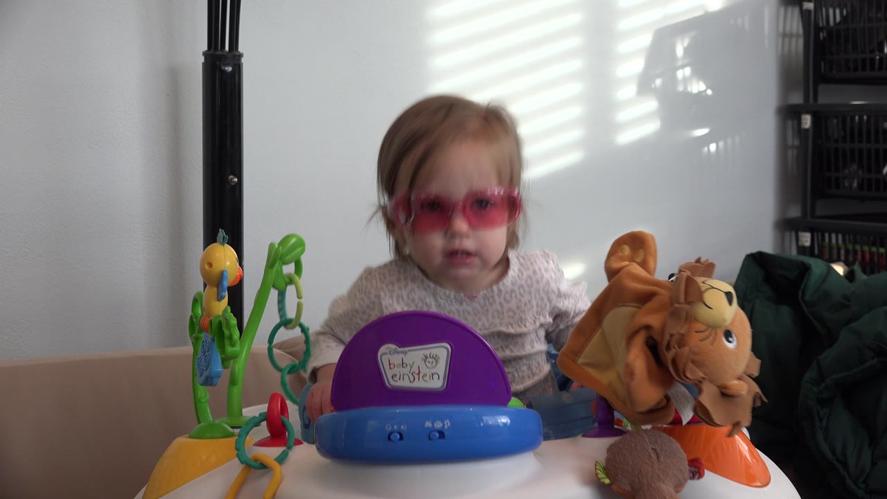 Baby in pink goggles sitting in a walker with colorful toys.