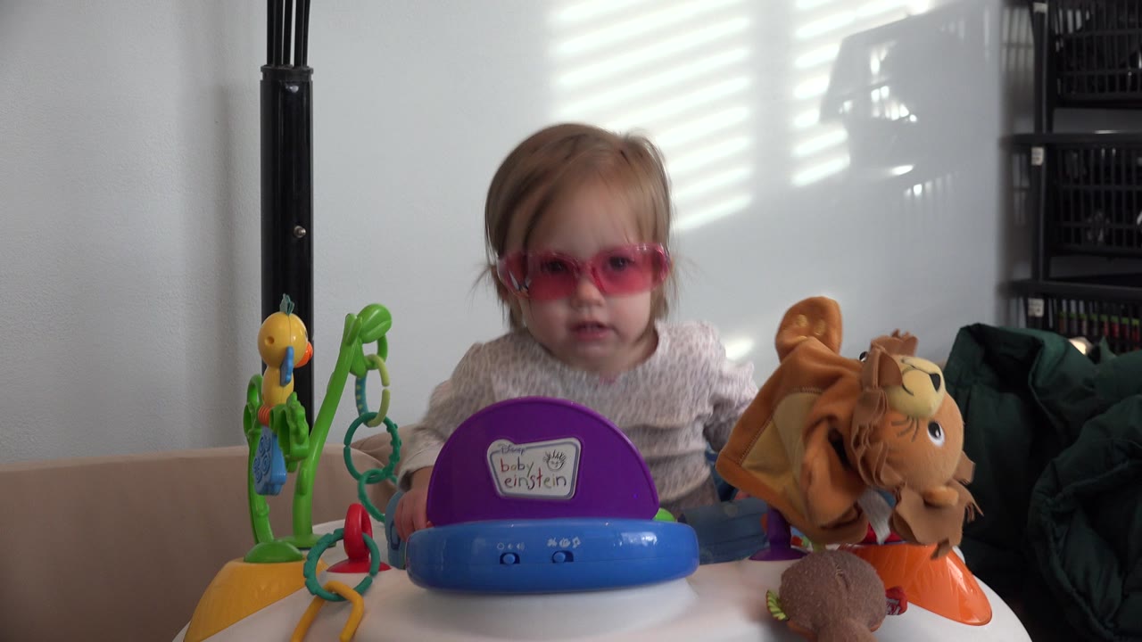 At 0:01 in Westminster, Colorado, the baby looks forward through pink goggles while seated behind a blue walker tray with a plush horse toy to the right.
