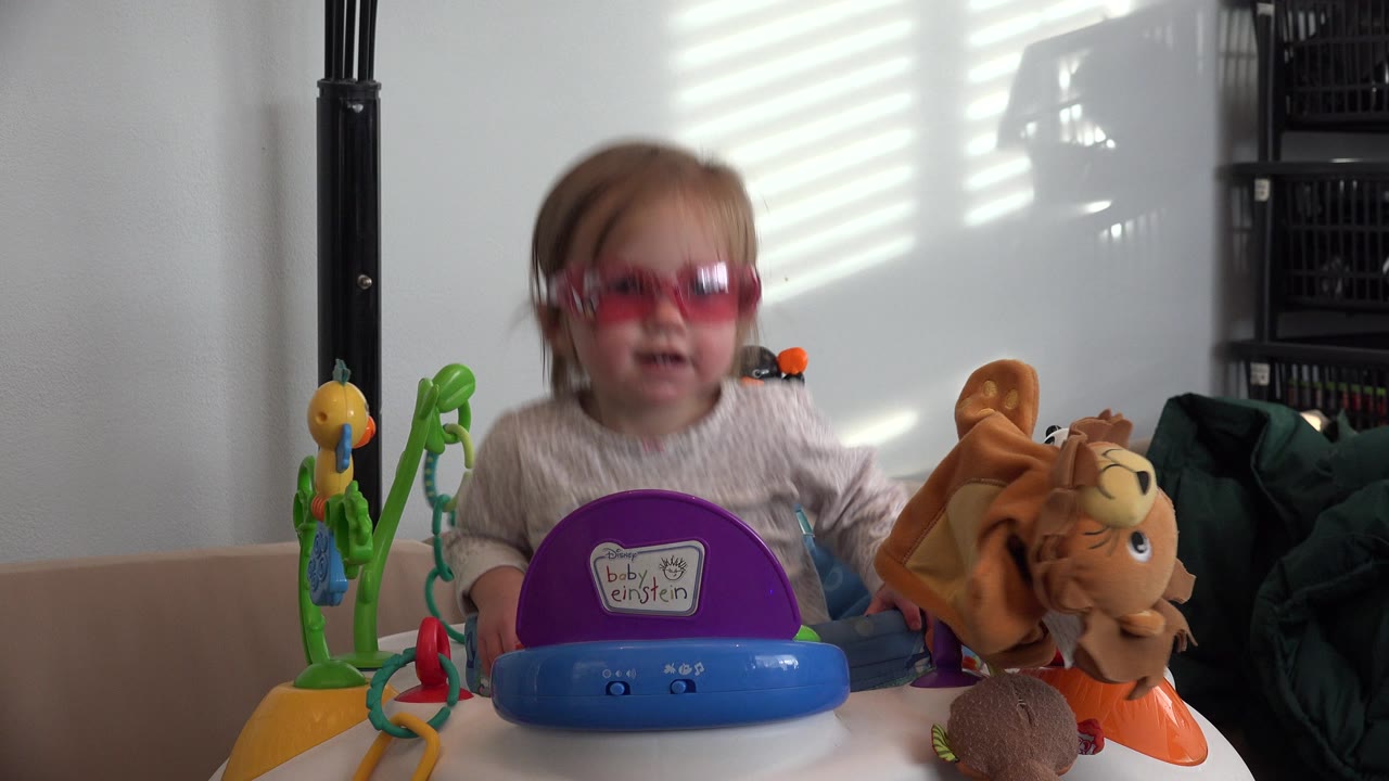 The toddler remains near the center of the Westminster, Colorado indoor frame, with a purple-and-blue toy in front and a plush animal to the right.
