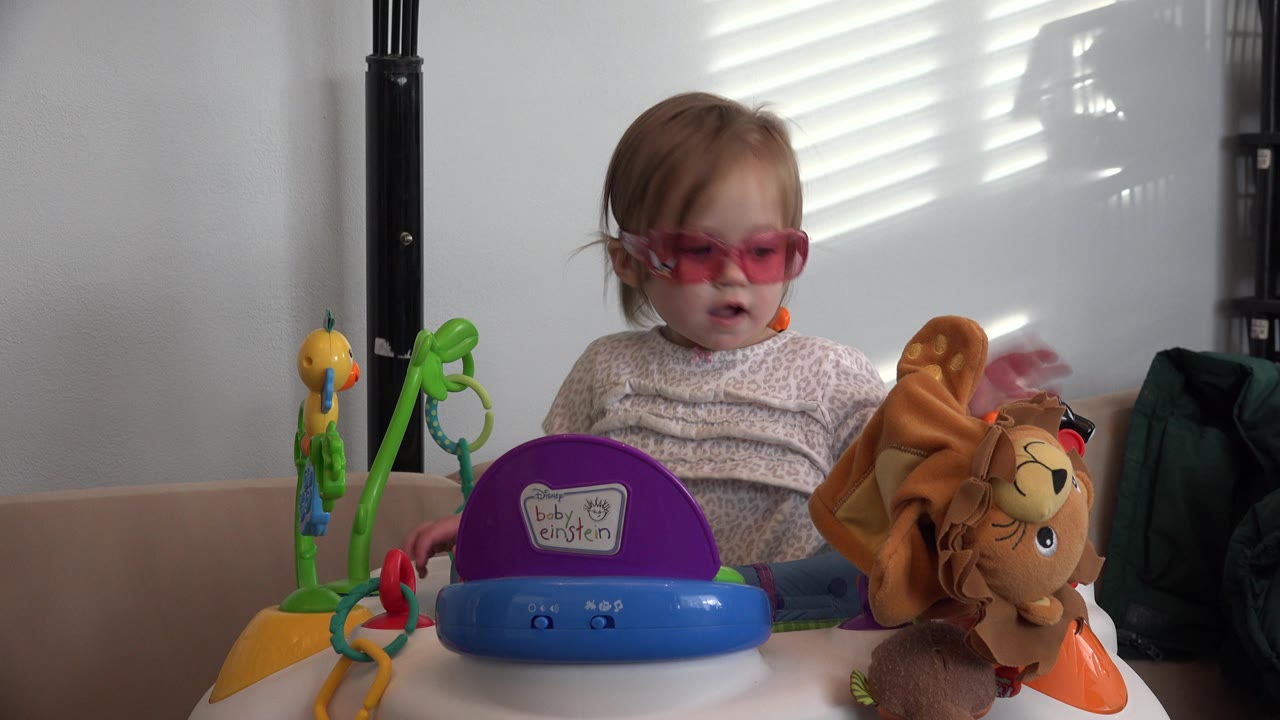 The child remains seated behind the colorful toy tray in Westminster, Colorado, facing slightly toward the plush lion as bright window-blind streaks fall across the wall.