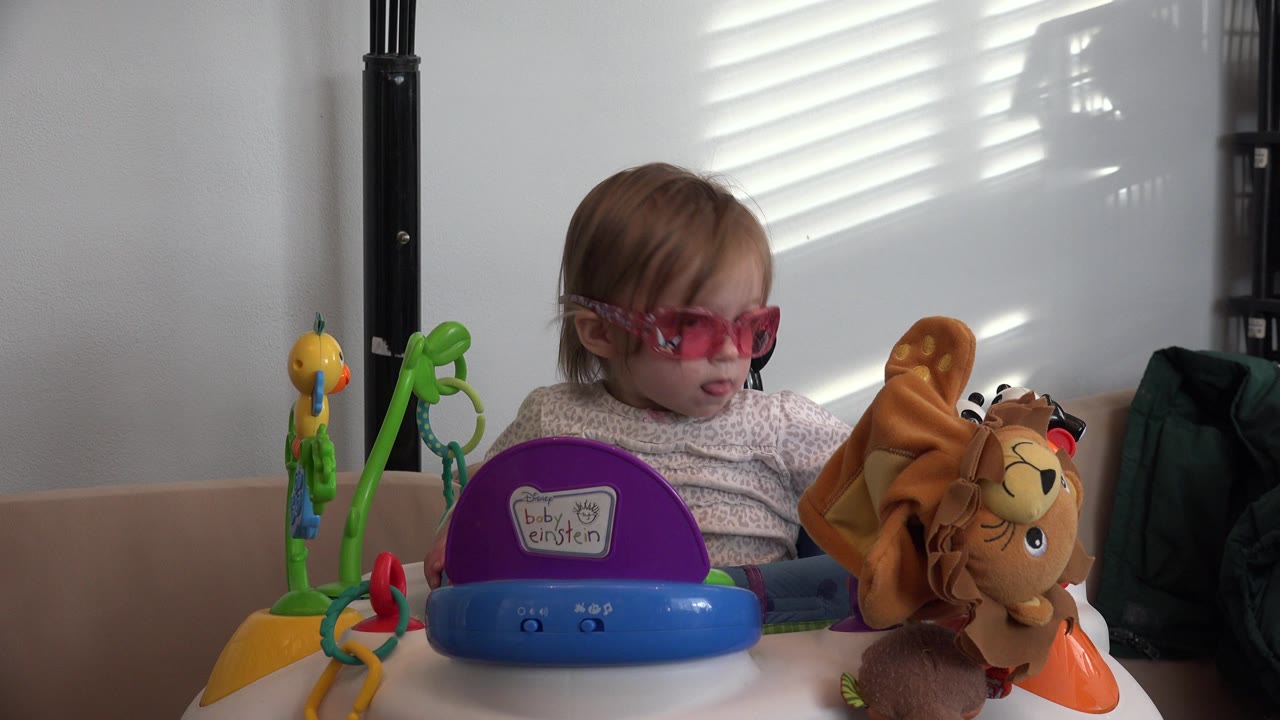 Toddler in pink glasses sitting behind colorful toys indoors.
