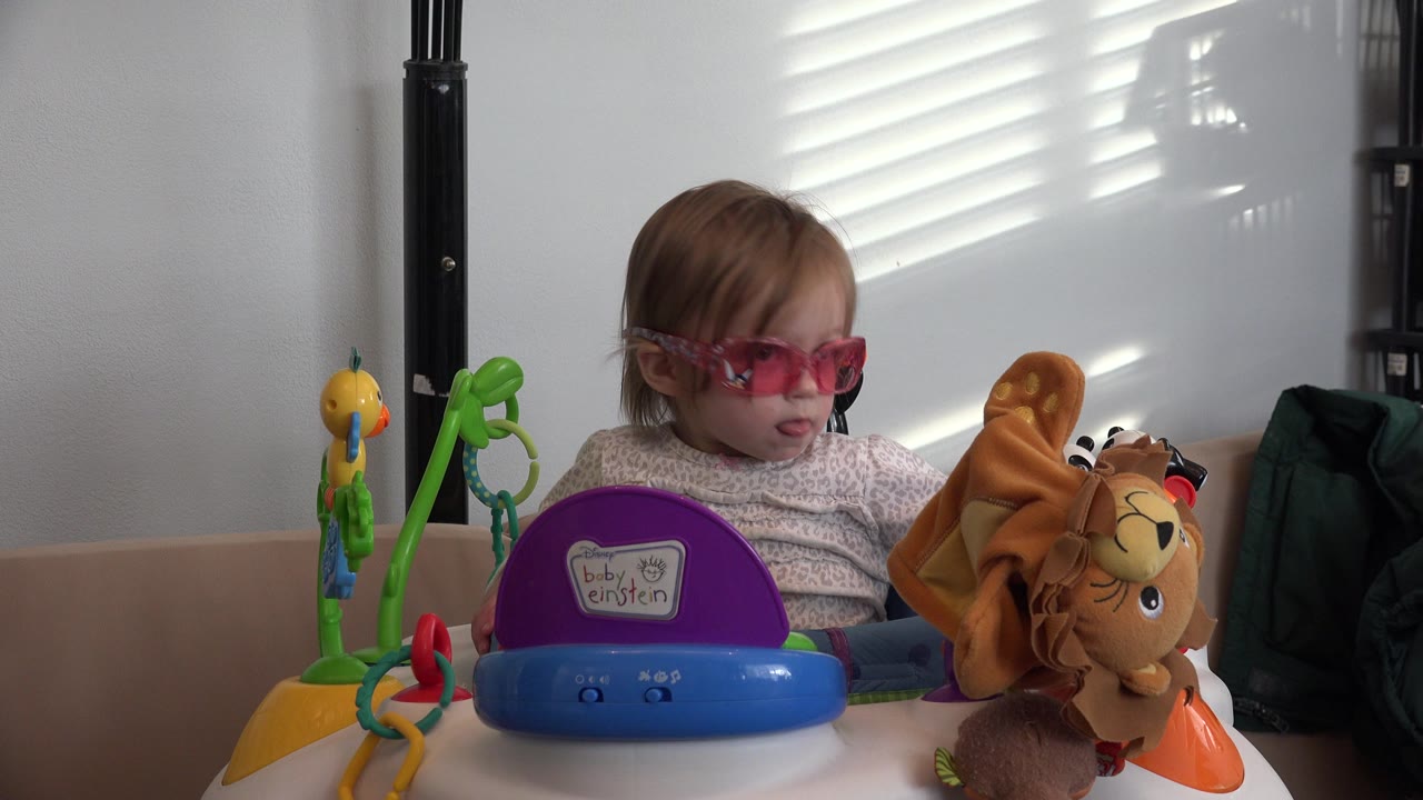 At 0:01, the child keeps their gaze lowered behind the purple Baby Einstein toy while the plush animal remains close to the right side of the frame.