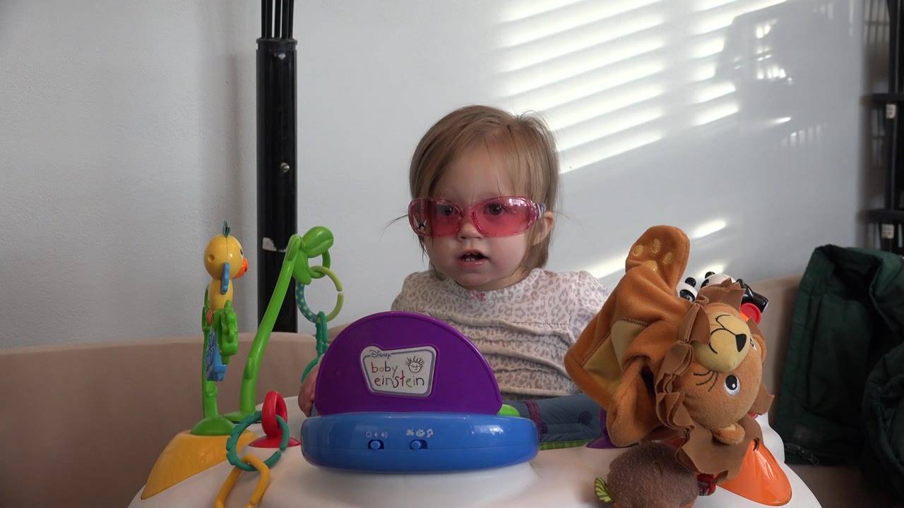 Child in pink sunglasses sits behind colorful toys at a table.