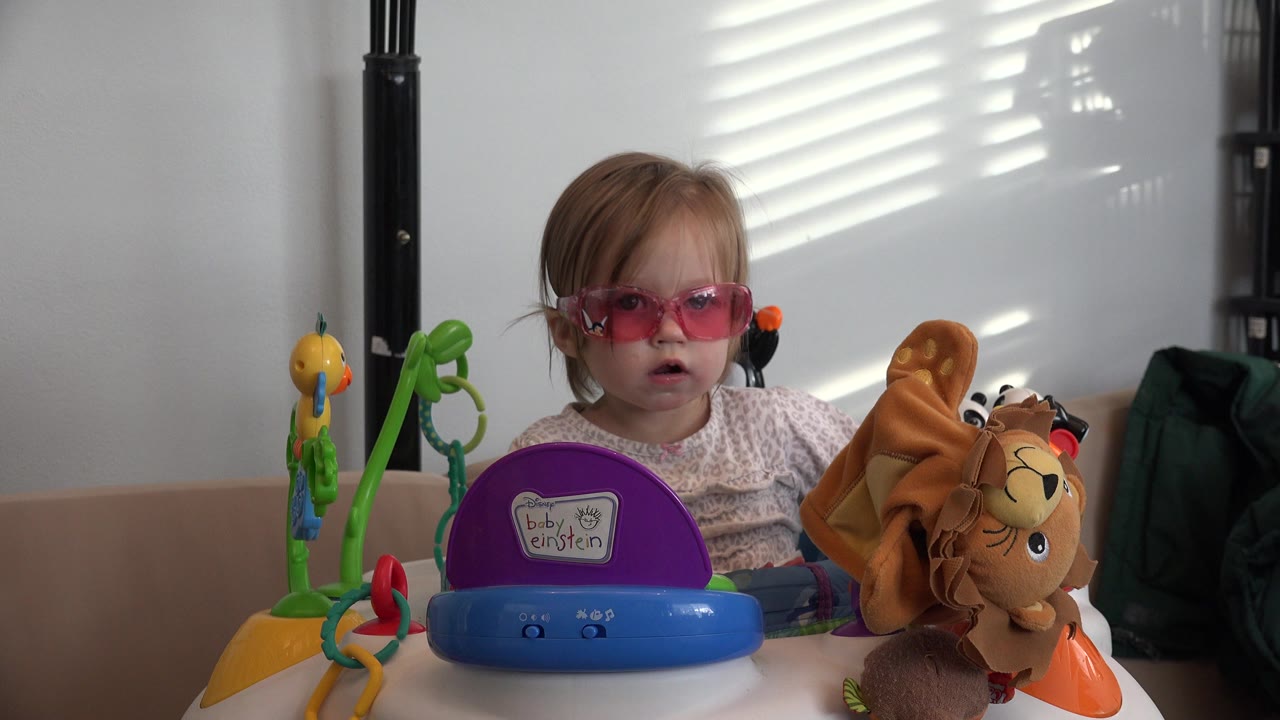 Little girl wearing pink sunglasses sits in a baby exerciser with toys.