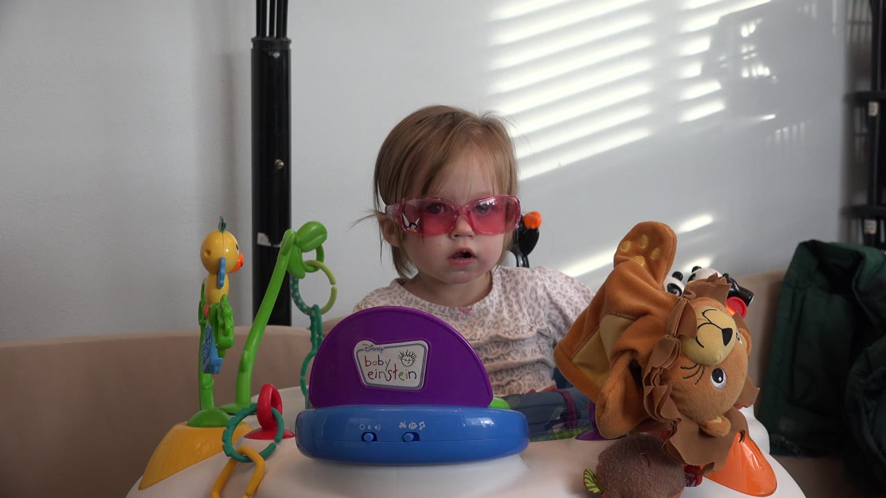 The child remains centered in the Westminster home video frame, facing forward through pink sunglasses while toys and striped window light surround the exerciser.
