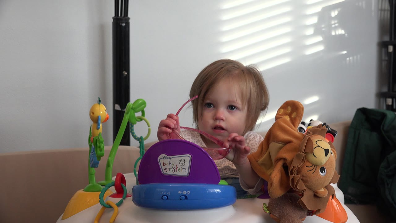 Blonde child behind colorful toys holding pink sunglasses indoors.