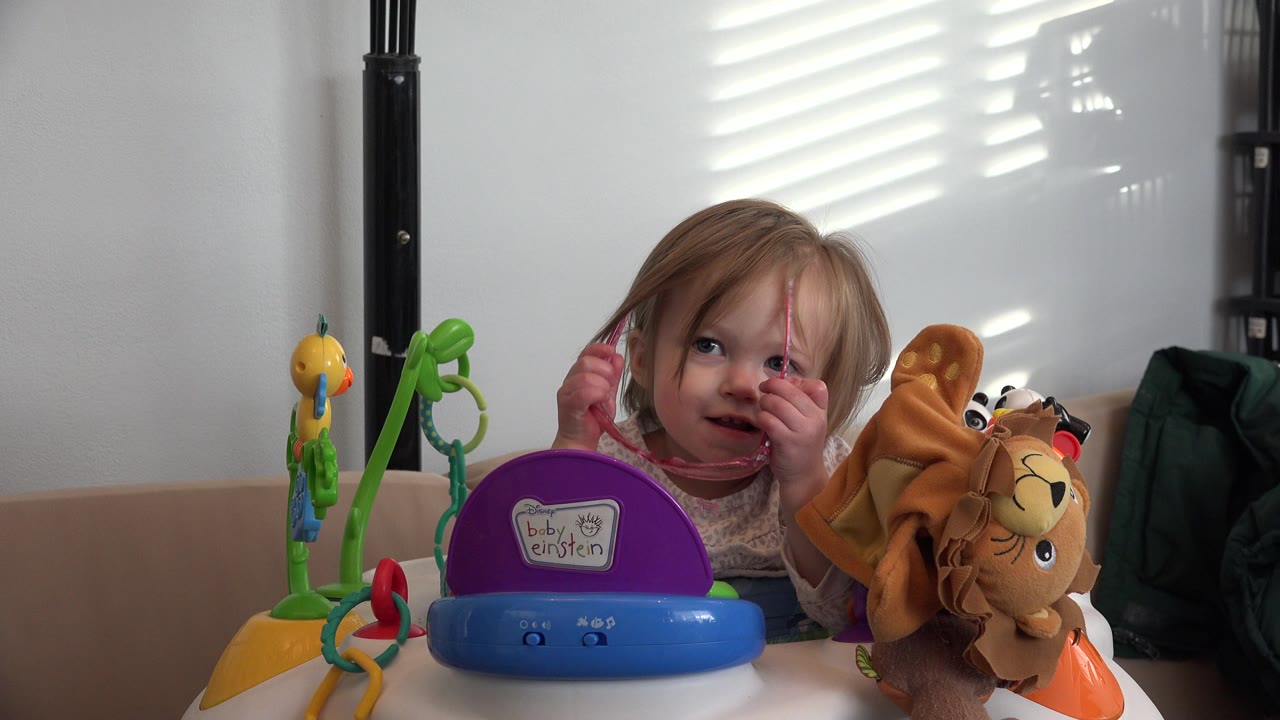 Blond baby girl holding pink glasses behind colorful activity toys.