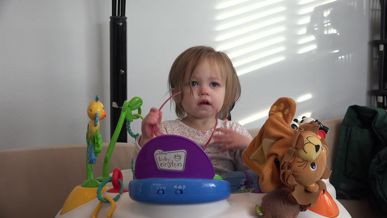 The toddler in Westminster, Colorado holds a pink cord near her face while seated behind the purple and blue toy with bright slatted light on the wall.