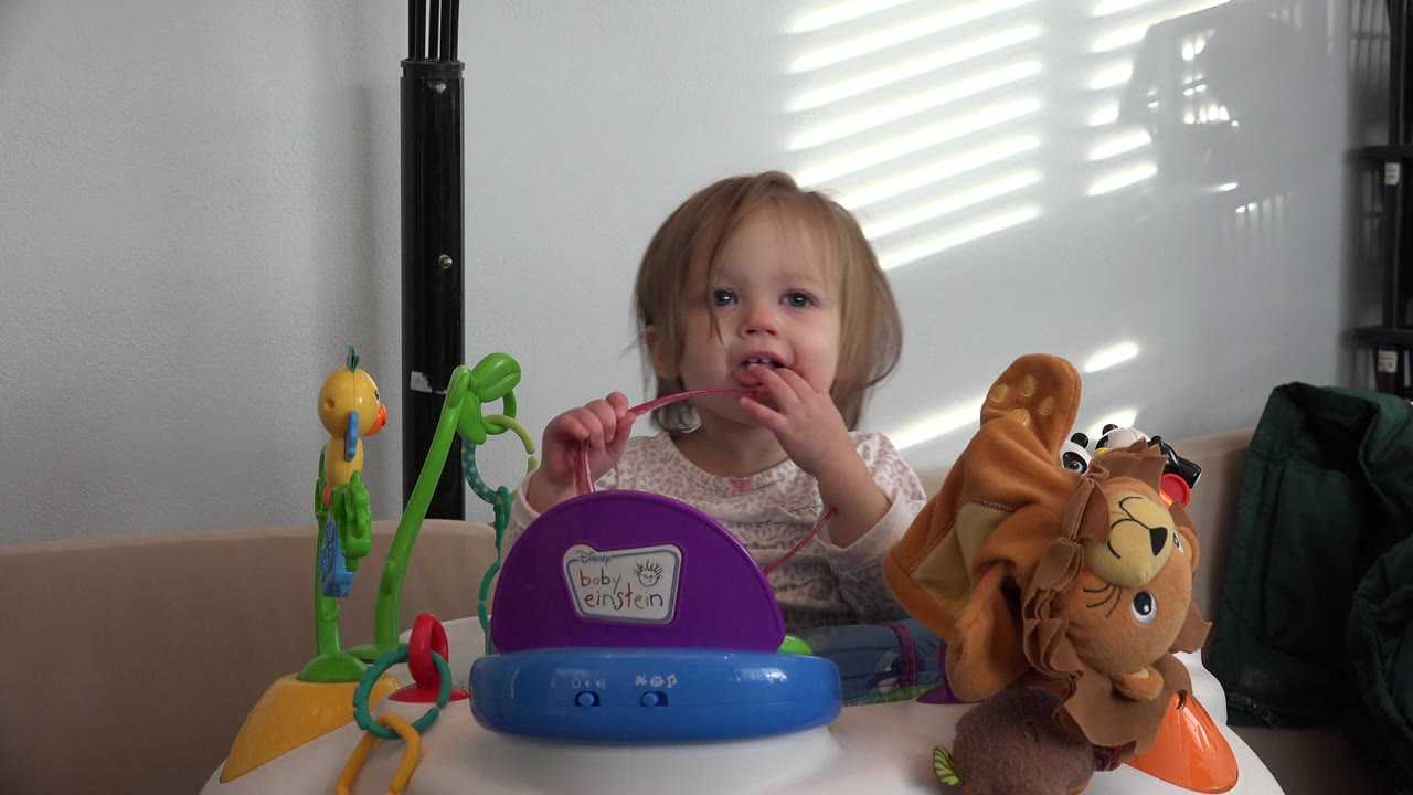 Baby in a walker holds pink glasses near their mouth beside toys.