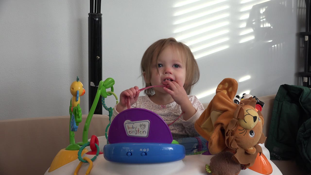 A baby in Westminster, Colorado looks forward while seated in a walker, holding pink glasses beside a purple tray and plush lion toy.